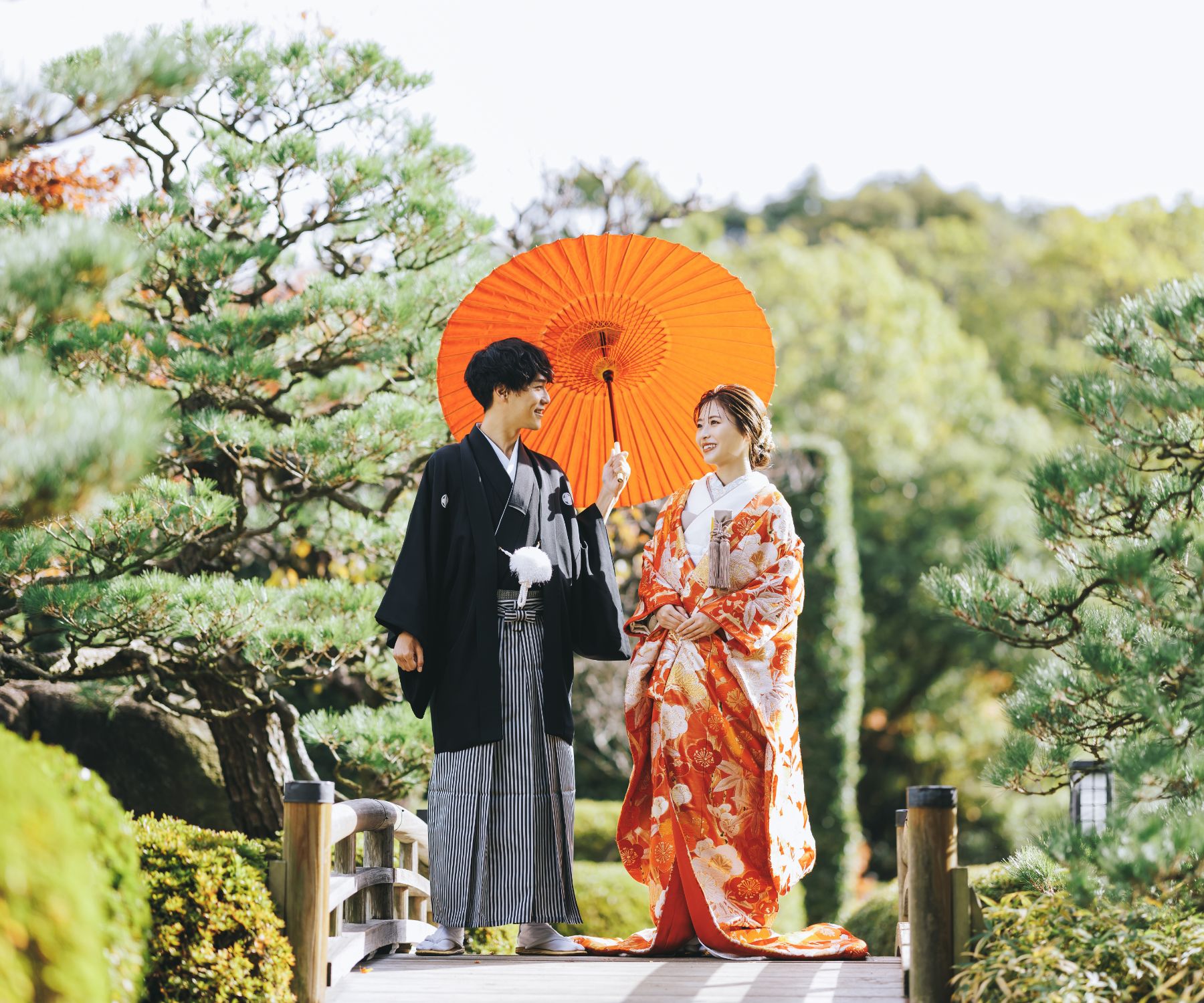 橋の上で赤い和傘をさして微笑む新郎様と新婦様の和装前撮り