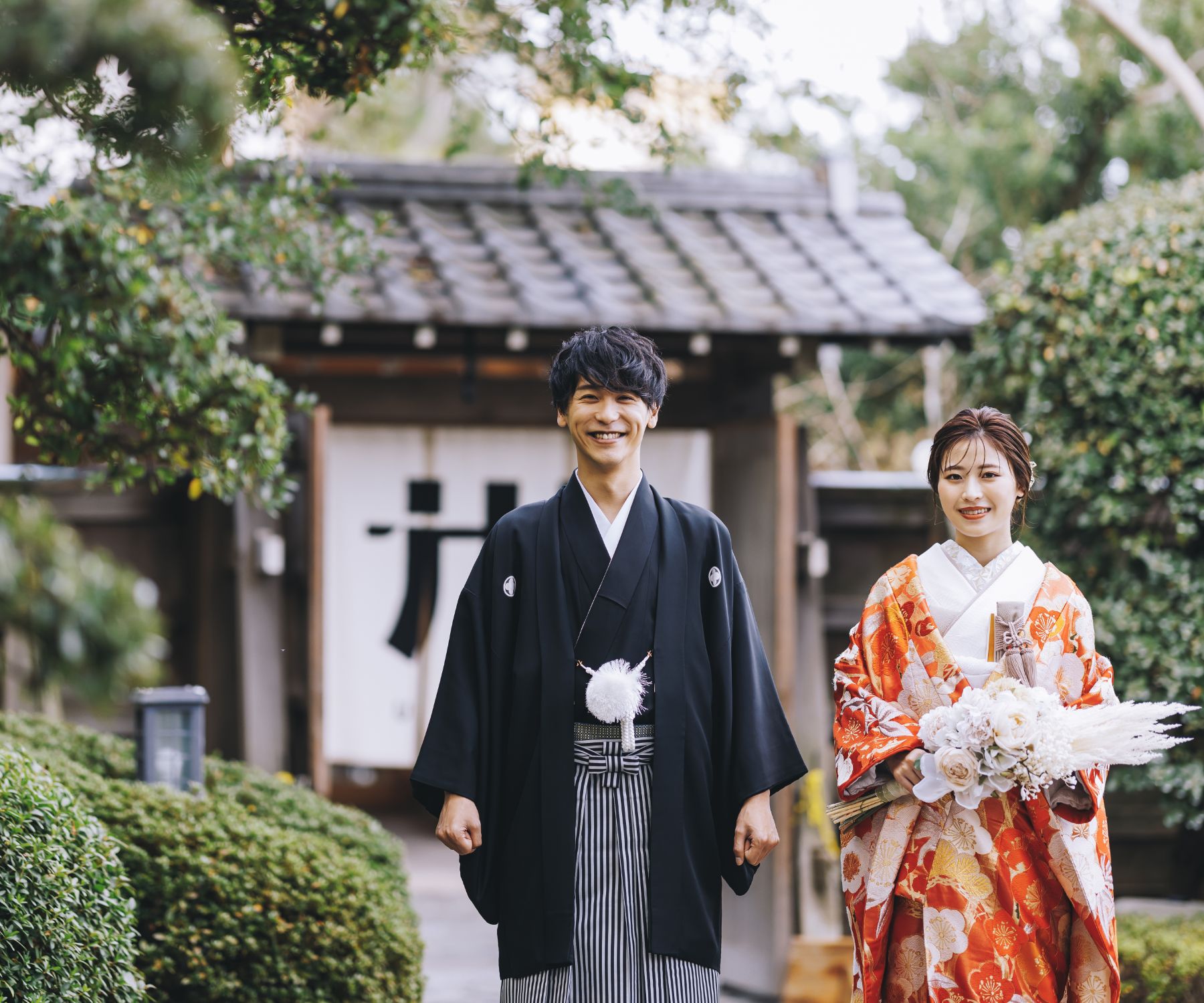 立派な門を背景に微笑む新郎様と花束を持った新婦様のロケーションフォト