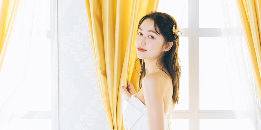                     光があふれる窓をバックにカーテンに触れながら微笑む新婦様