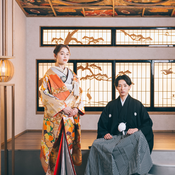                和柄の背景の前でポーズをとるタキシードを着た新郎様と色打掛を着た新婦様の結婚写真     