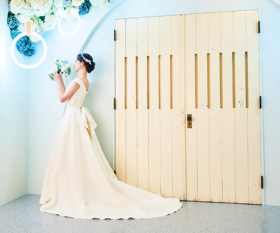      淡いブルーの背景セットで花束を抱える新婦様のバックショット               